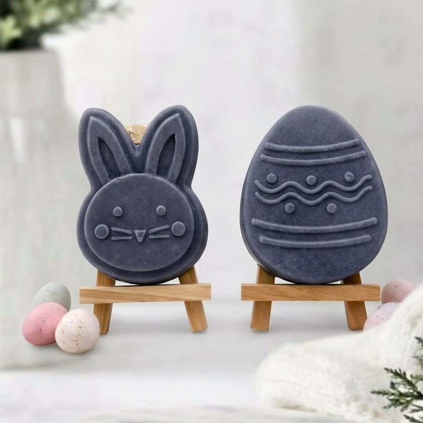 Sophisticated dark charcoal Easter soap gift set for a luxury bath. Two novelty vegan soaps, a grey bunny face and a matching patterned egg, are presented on miniature wooden easels on a marble tray with speckled pastel miniature egg