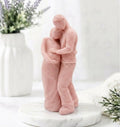 A handmade soft pink soap sculpture. It depicts a man in pants and a shirt embracing a pregnant woman in a long dress from behind, with his hands over her belly. They are on a round marble stand