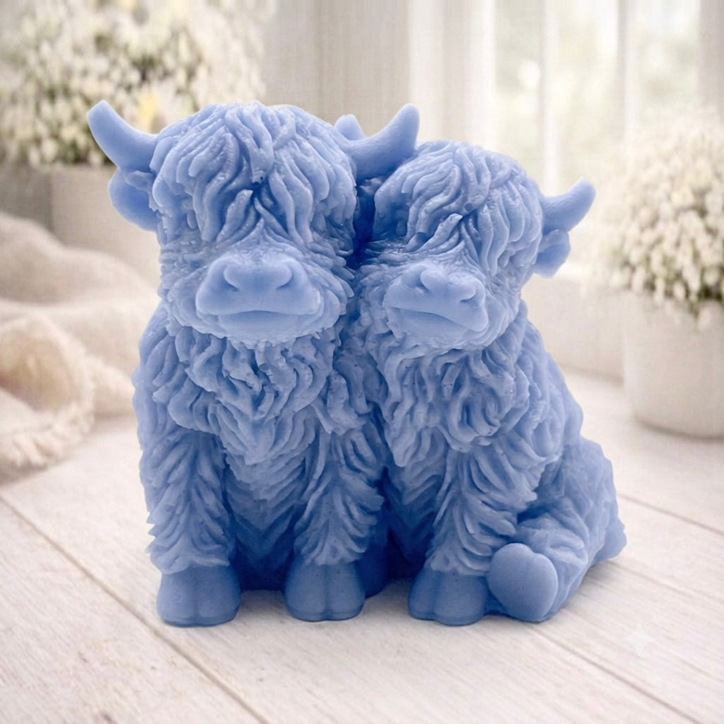 Handmade soap featuring two Highland cows with textured, shaggy fur sitting side by side on a light wooden surface with a softly indoor background.