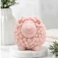 A pink sheep / ram soap with curly wool details, standing on a round white marble base against a soft, blurred background with a white vase and greenery.