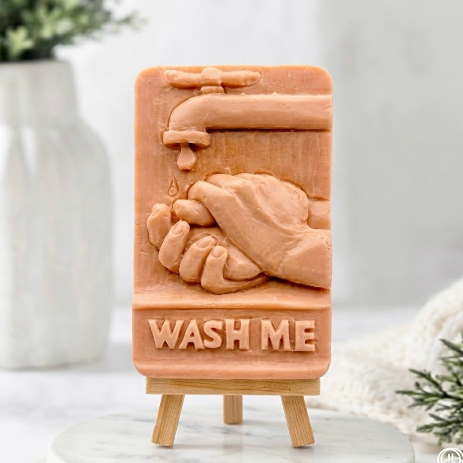 A terracotta-coloured sculpted vegan soap bar featuring a detailed relief carving of hands washing under a water tap and the text "WASH ME," presented on a miniature wooden display easel in a spa-like setting.