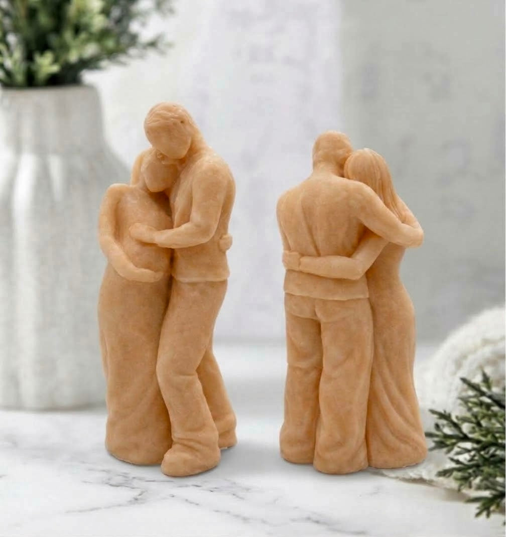 A front and rear view of a handmade soap sculpture. It depicts a man in pants and a shirt embracing a pregnant woman in a long dress from behind, with his hands over her belly. They are on a round marble stand