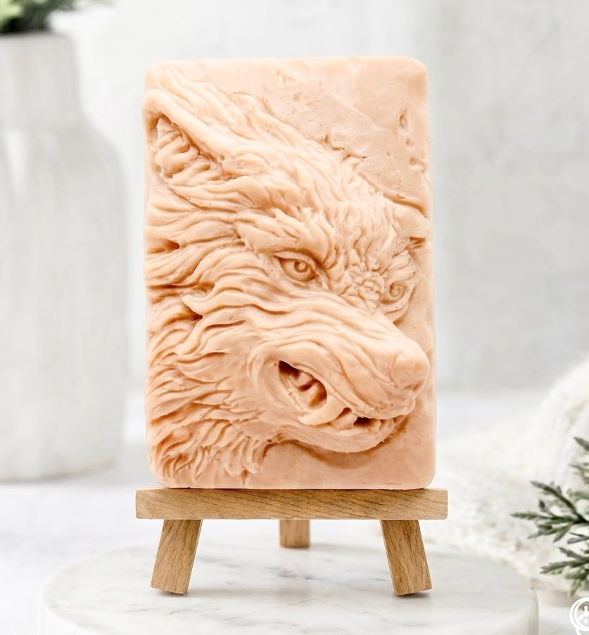 A rectangular pink French clay soap bar featuring a detailed relief carving of a wolf's head, displayed on a small wooden easel stand against a soft, light gray and white background.