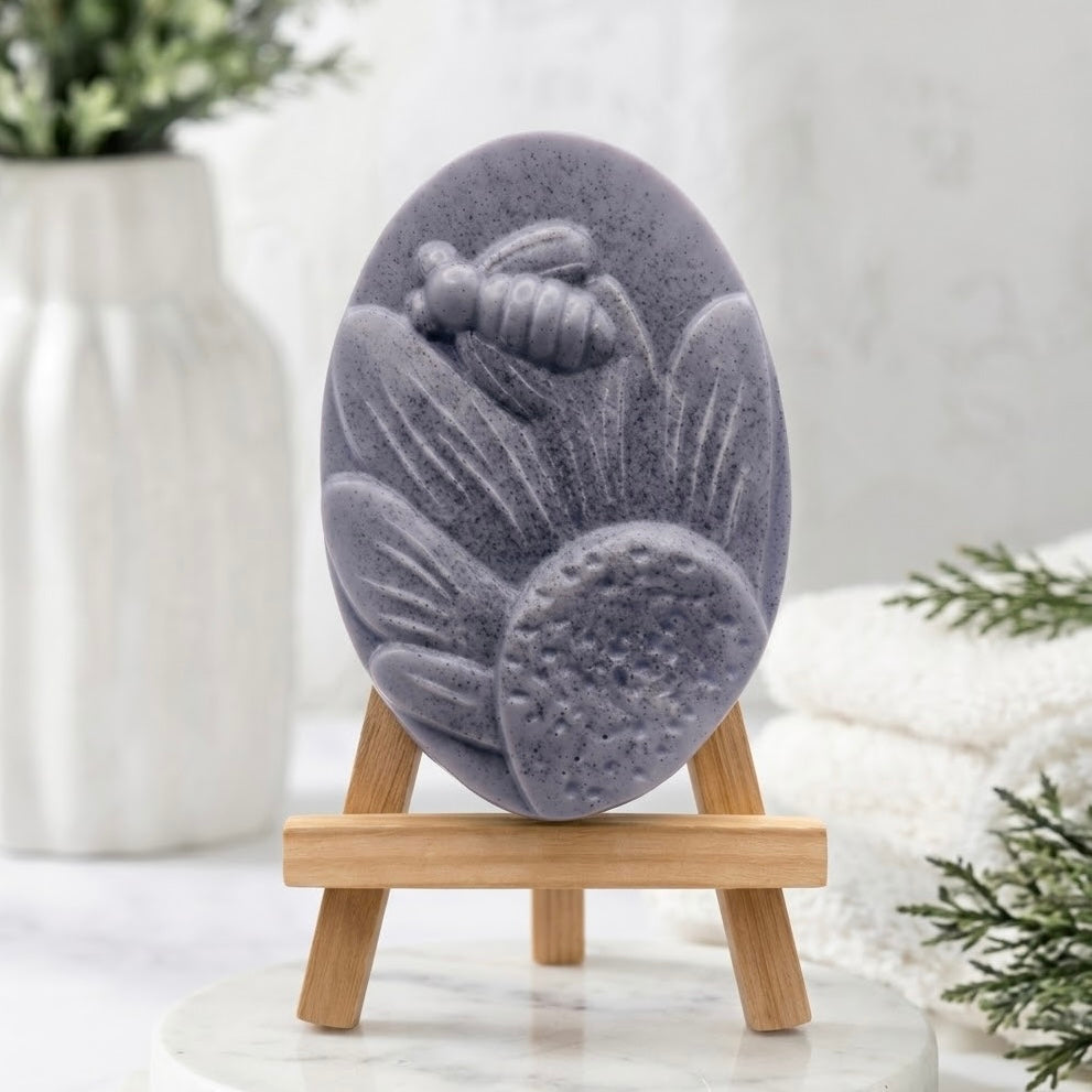 A handmade vegan soap in a charcoal grey colour, featuring an oval relief of a honeybee landing on a large sunflower with detailed petals and a textured centre, displayed on a small wooden easel.