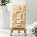 A delicate, soft yellow artisanal soap, standing vertically on a small wooden easel. The soap features a detailed relief carving of a large butterfly with two smaller ones fluttering among swirling floral and leaf motifs. It's displayed on a marble base with a blurred white vase and soft greenery in the background.