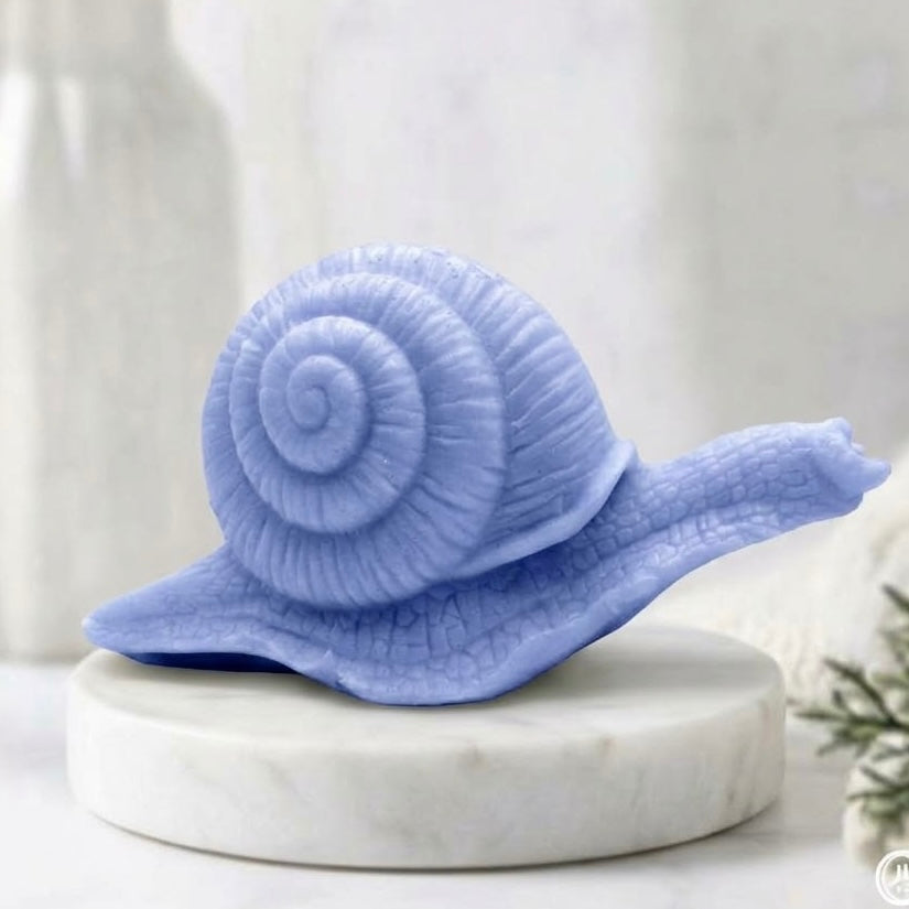 A highly detailed handmade vegan soap in a soft blue colour, sculpted in the shape of a realistic snail with a prominent spiralled shell and a textured, leathery body, displayed on a light wooden surface.