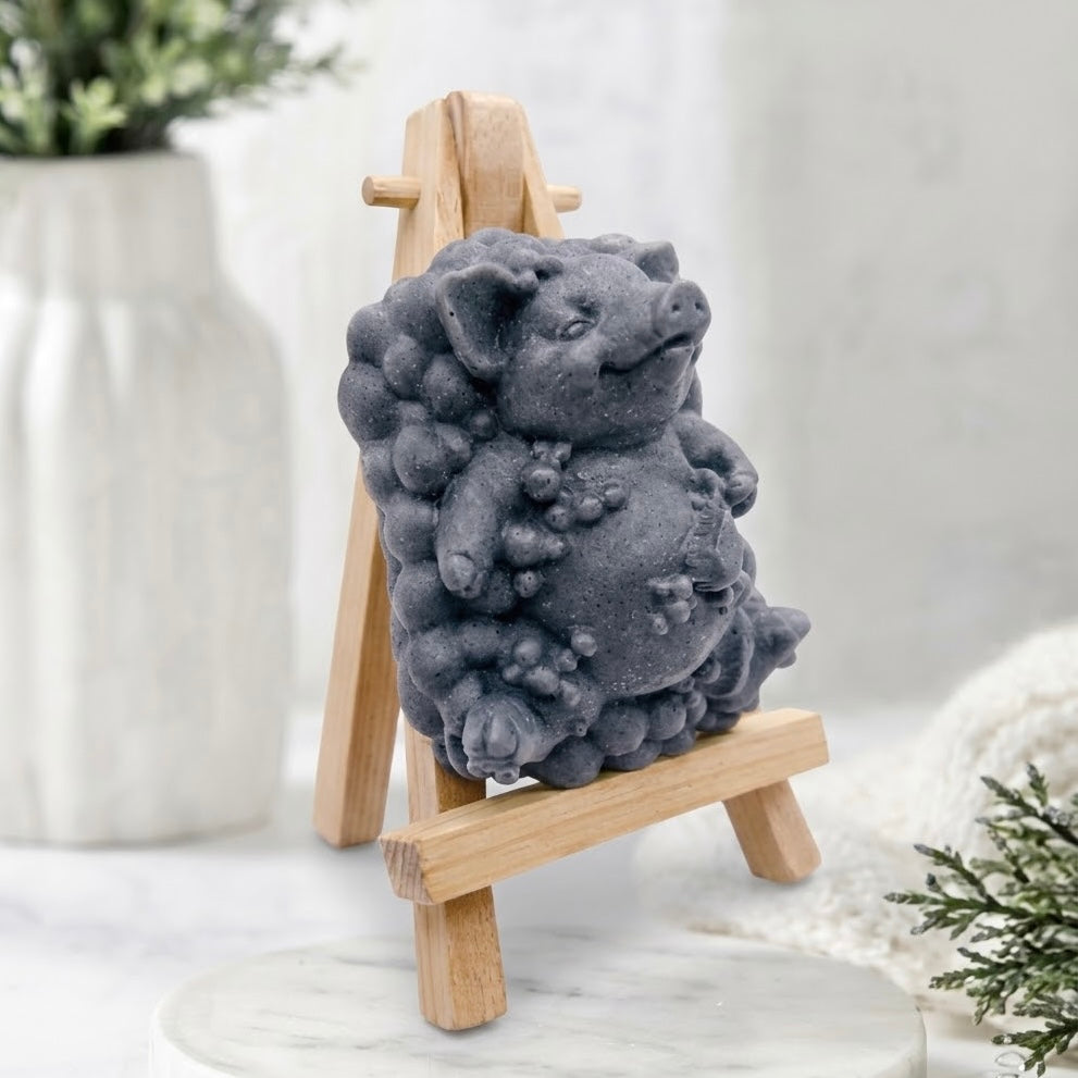 A three-quarter angle view of a deep charcoal grey pig-shaped soap figurine with a bubble-cluster base, showcasing the dark colour and intricate details, resting on a miniature wooden easel.