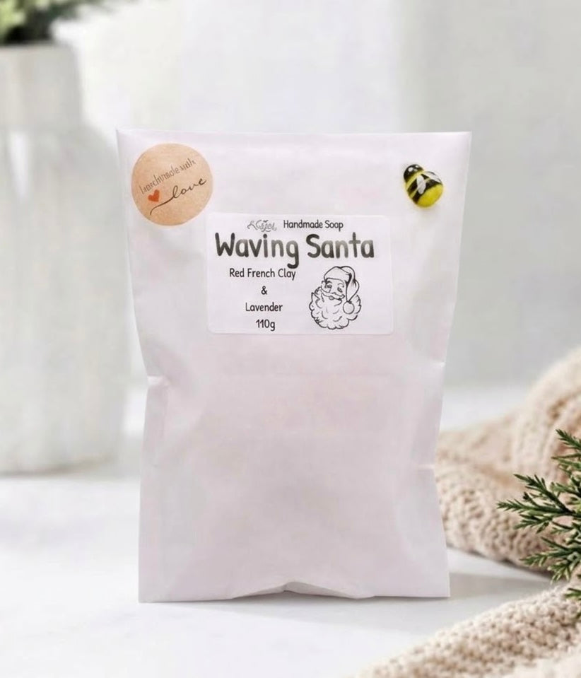 A photograph of a clean white paper gift bag with multiple labels. The main label features text that reads 'Handmade Soap' and 'Waving Santa', with 'Red French Clay & Lavender' and '110g' beneath a charming illustration of Santa Claus. A round brown sticker at the corner says 'handmade with love' and has a small heart, and a small, three-dimensional bumblebee is clipped to the corner.