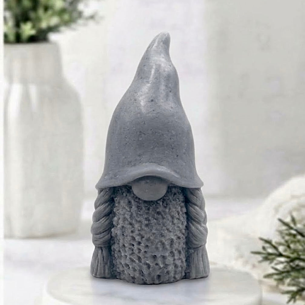 A handmade charcoal grey vegan soap in the shape of a female gnome (or gonk), featuring a tall, slightly curved hat pulled over the eyes and two thick, textured plaits framing a textured dress.