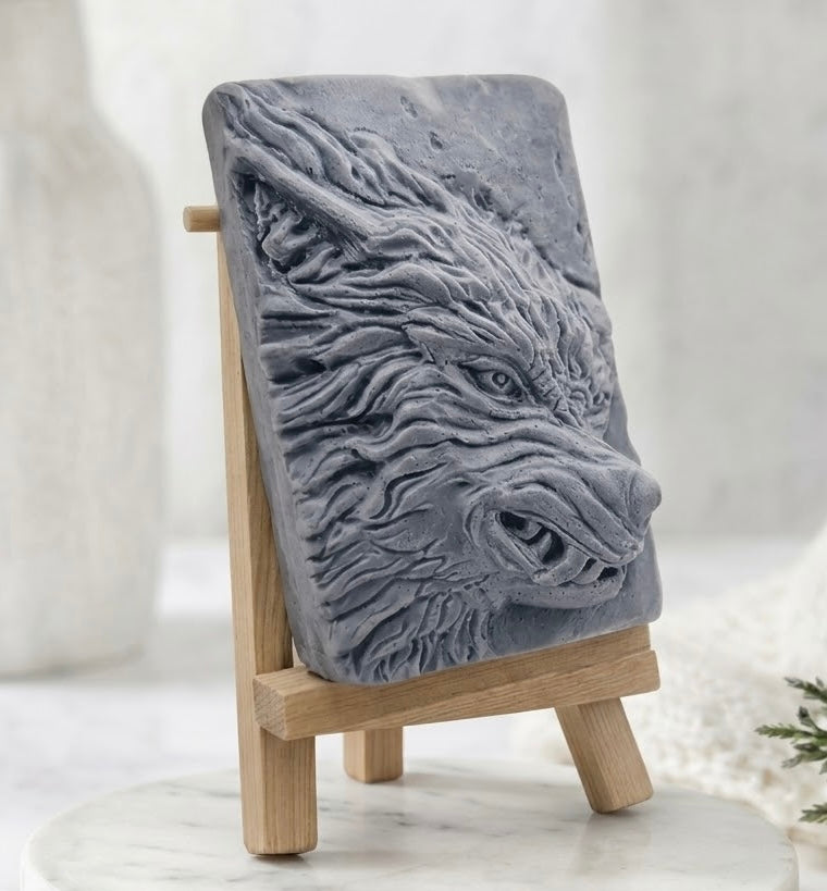 A charcoal gray vegan soap featuring a detailed relief carving of a wolf's head, displayed on a small wooden easel stand against a soft, light gray background.