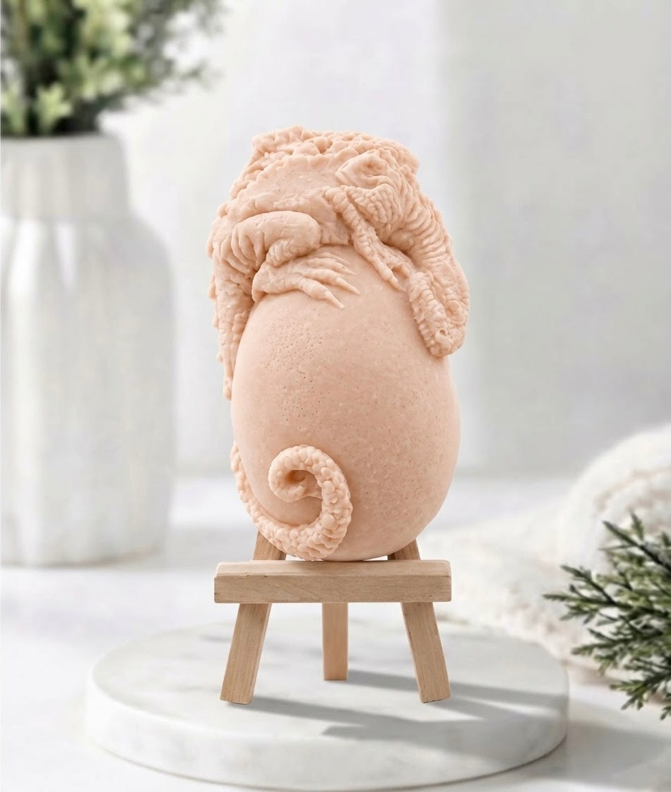 A pink French clay vegan soap resembling a stylised egg abs dragon with textured, curly details, displayed on a small wooden stand on a white marble-like surface. 