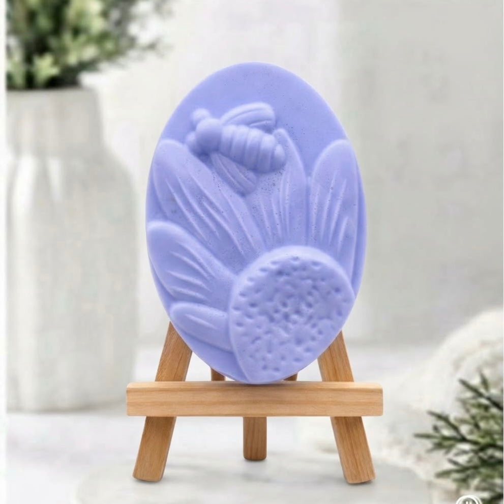A handmade vegan soap in a soft blue, featuring an oval relief of a honeybee landing on a large sunflower with detailed petals and a textured centre, displayed on a small wooden easel.