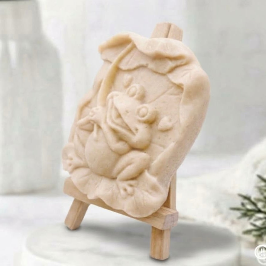 A side view of a handmade novelty frog vegan soap bar in a soft yellow colour, presented in a unique animal design on a miniature wooden display easel.