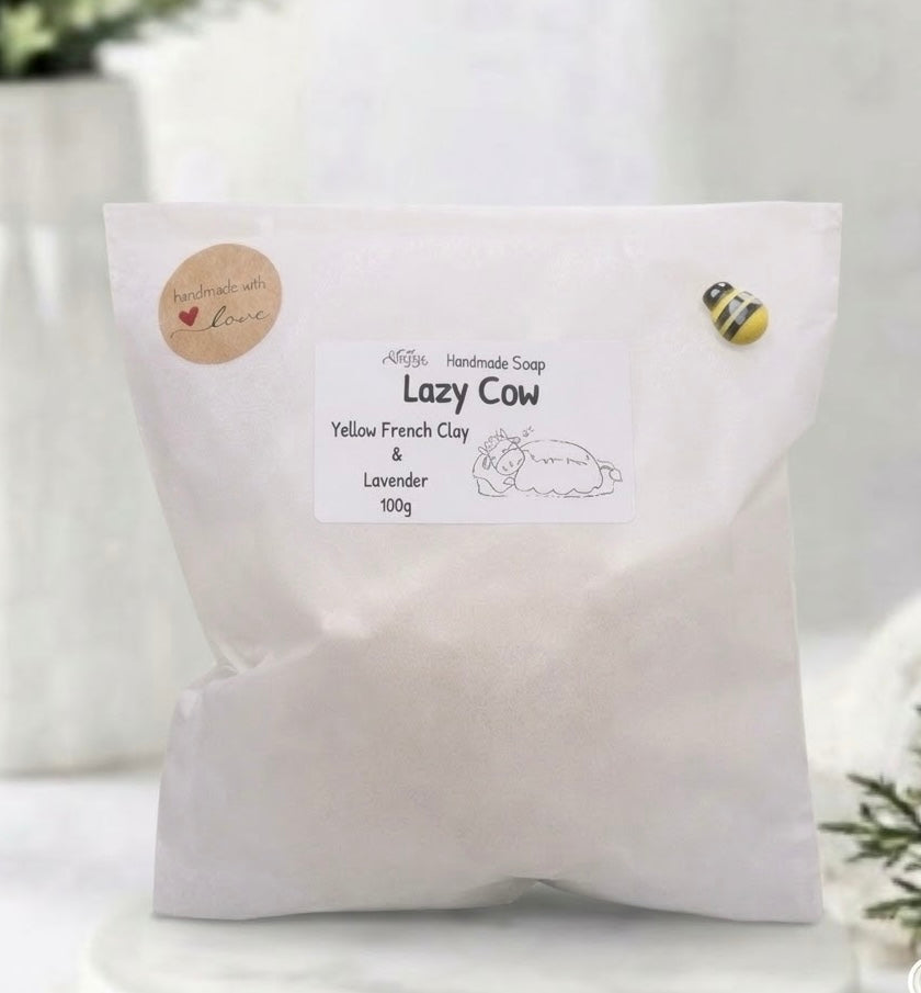 A white paper bag of Lazy Cow handmade soap by The Happy Handmade Co, featuring a white label with black text and a small bee magnet on the bag, set against a soft, blurred white background.