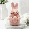 A pink bunny-shaped decorative gnome soap with long ears, a small nose, and a textured body, standing on a round white marble base against a soft white background with blurred greenery and fabric.