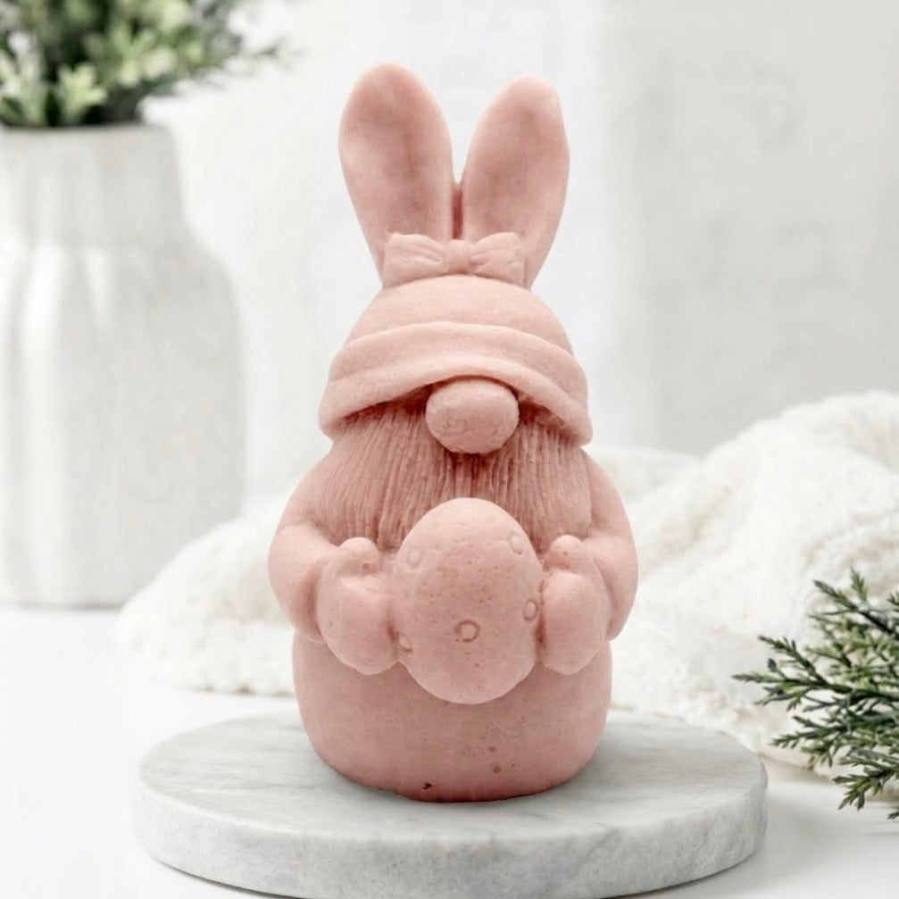 A pink bunny-shaped decorative gnome soap with long ears, a small nose, and a textured body, standing on a round white marble base against a soft white background with blurred greenery and fabric.