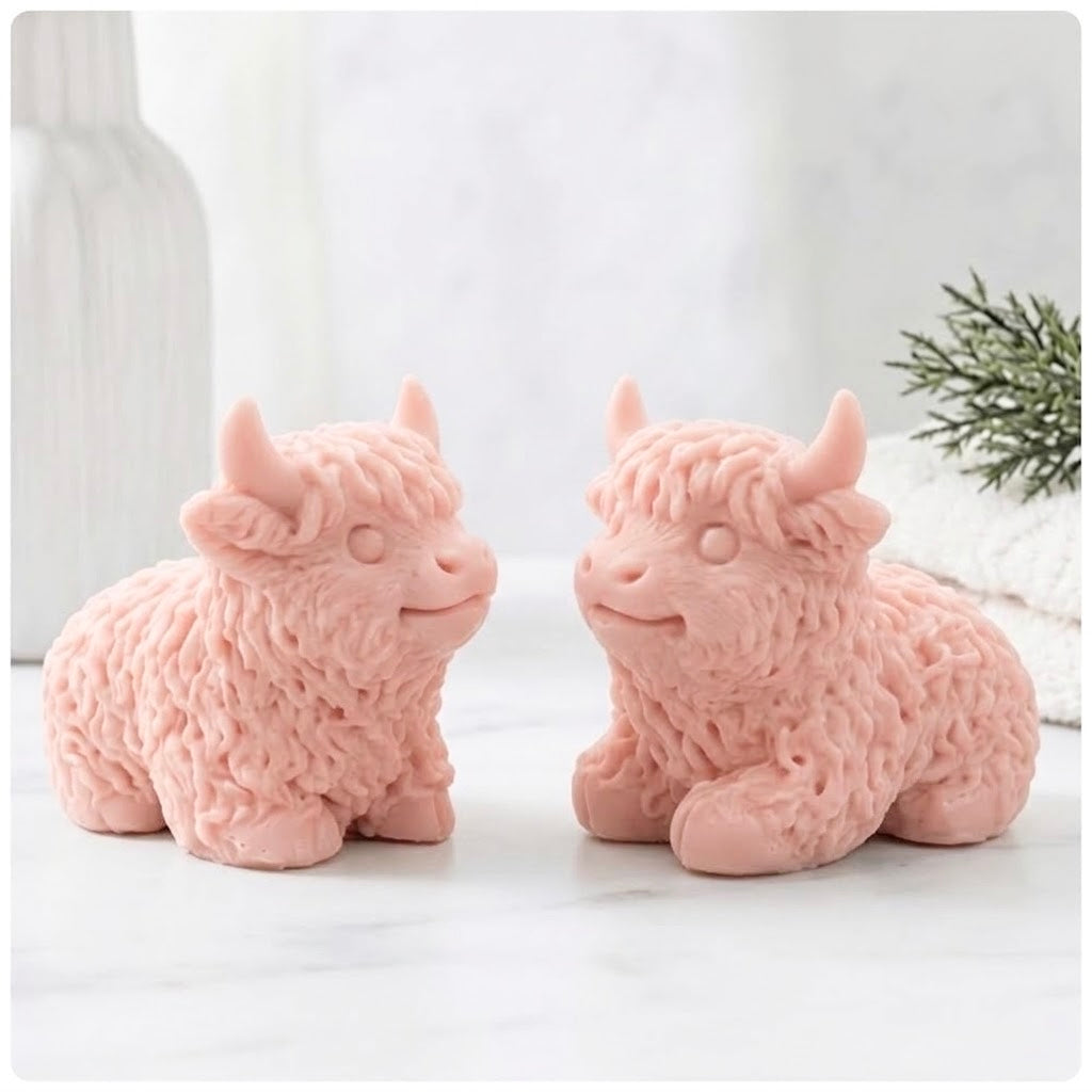 Two pink French clay Highland cow soaps are sitting side-by-side and facing each other, on a surface with a blurry background of a vase and a towel.