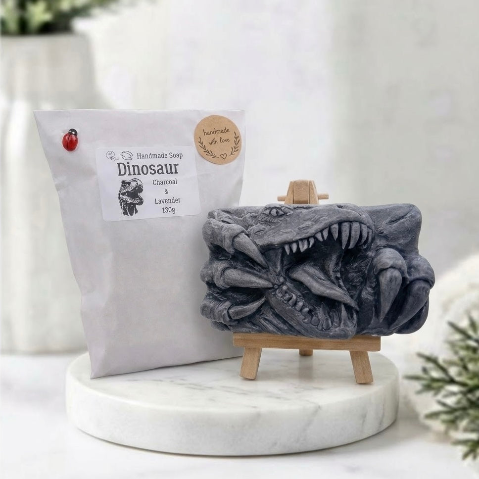 A rectangular grey handmade vegan soap with packaging, featuring a high-relief carving of a Tyrannosaurus Rex with its jaws wide open in a roar and sharp claws framing the face, displayed on a miniature wooden easel.