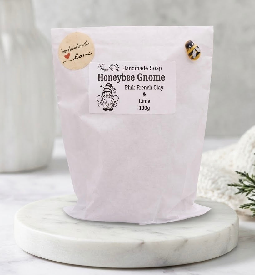 A photograph of the standing white paper packaging bag for the "Honeybee Gnome Pink French Clay & Lime" handmade soap, centered on a large round white marble pedestal against a blurred bathroom background.