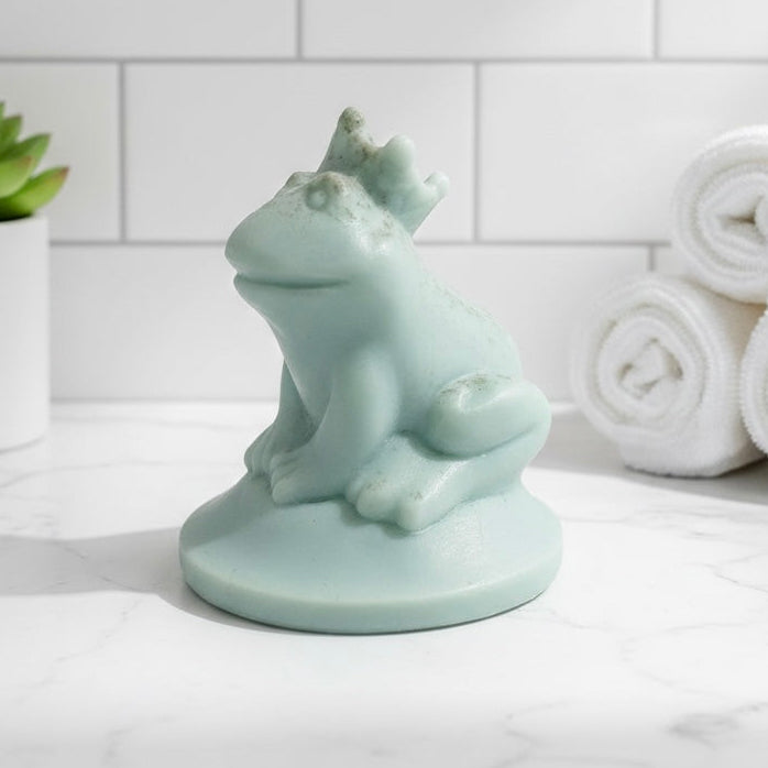 A 3D handmade vegan soap in a pale mint-green tone, sculpted into a sitting frog wearing a crown on its head, positioned on a circular base and surrounded by delicate white flowers in a bright farmhouse setting.