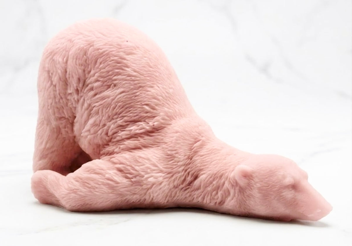 A light pink rose-colored bar of soap shaped into a detailed lazy polar bear figure lying down, resting on a white marble surface.