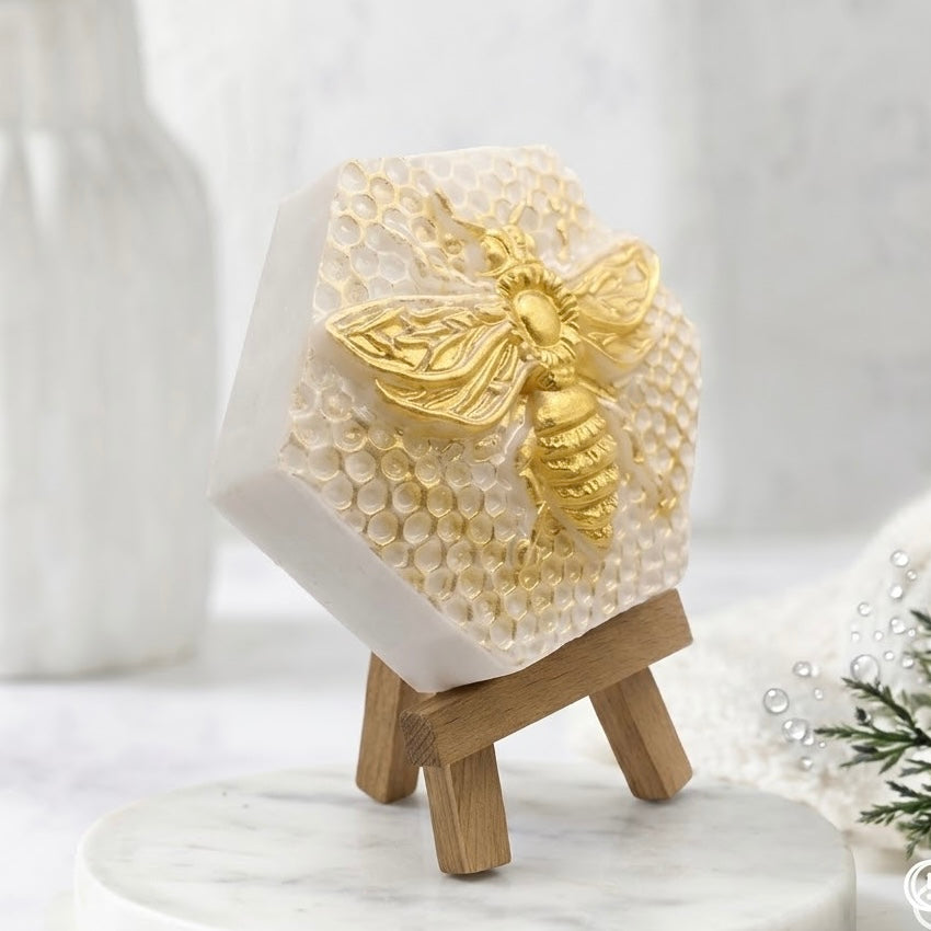 A closer three-quarter view of the hexagonal honeycomb soap from the right, emphasizing the detailed relief and its depth. The gold bee and textured honeycomb pattern are sharp. The small wooden easel and marble plate are visible, along with the spa-like background elements.