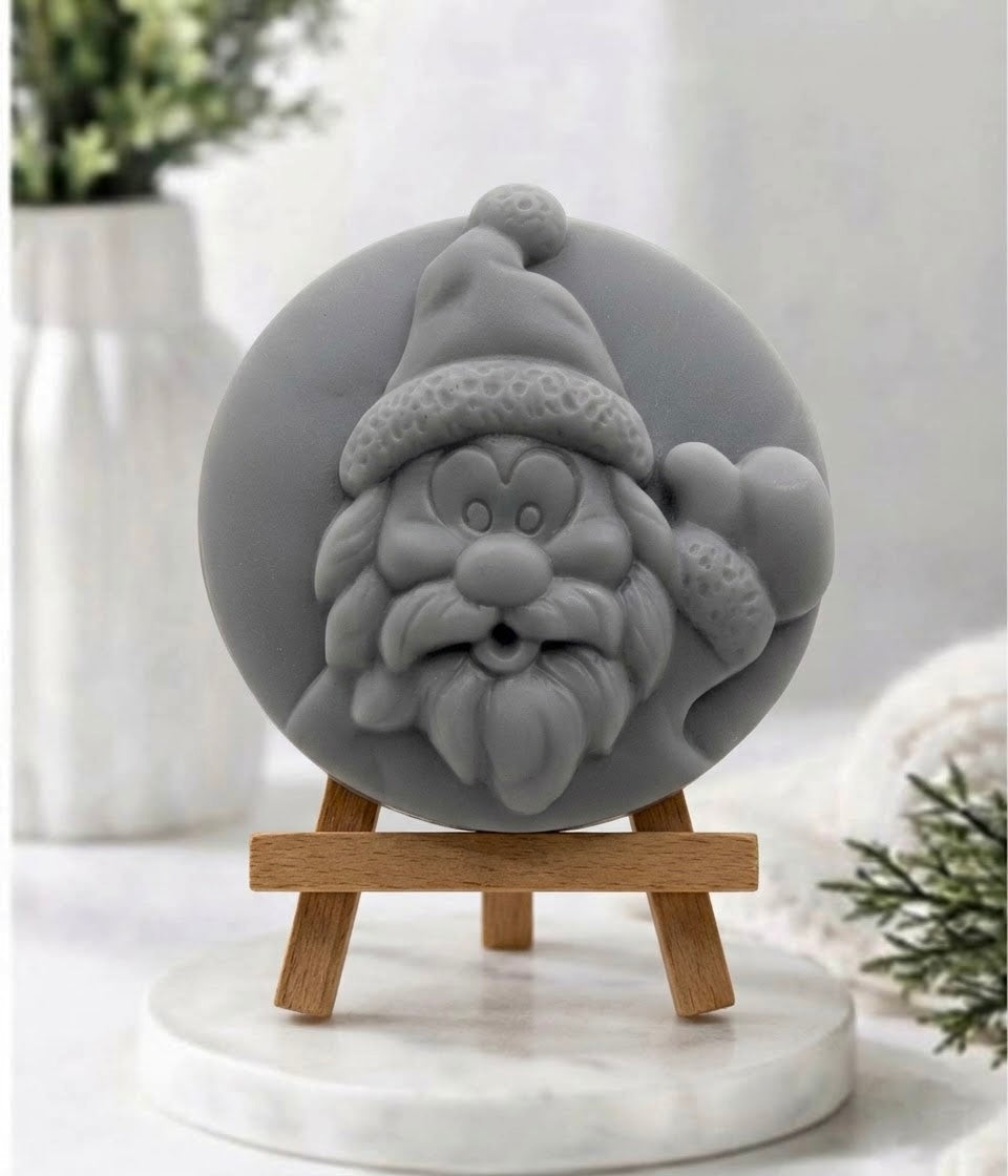 A detailed product shot of a circular, hand-carved soap disc with a relief of Santa Claus’s face, rendered in a solid, charcoal-grey colour. It is displayed on a small, light-wood tripod easel, which stands on a round white marble pedestal. The background features a soft-focus white vase, blurry greenery, and a cosy white textile.