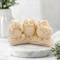 Three wise owl soap sitting on a branch-shaped base, displayed on a round white marble plinth in a softly lit indoor setting.