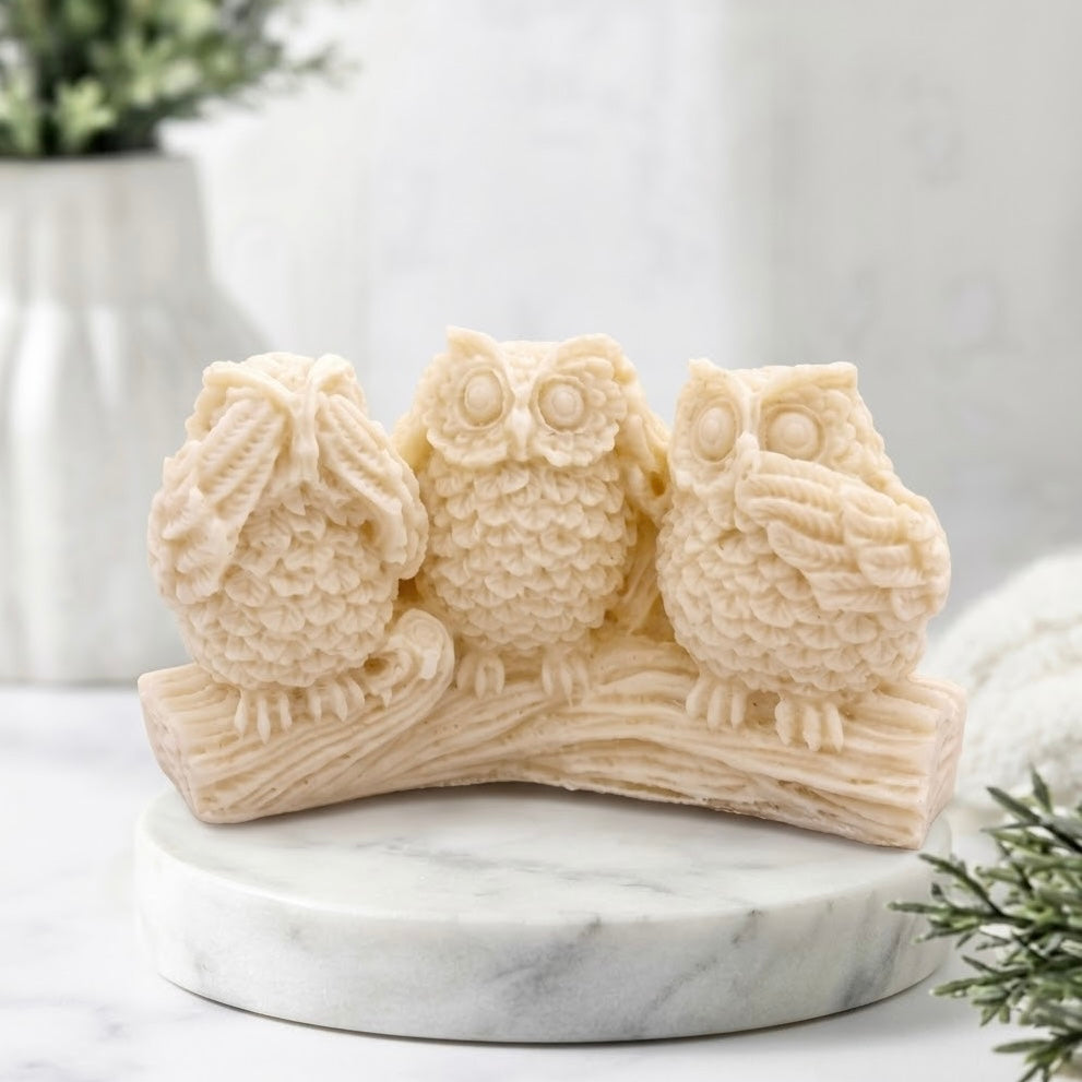 Three wise owl soap sitting on a branch-shaped base, displayed on a round white marble plinth in a softly lit indoor setting.