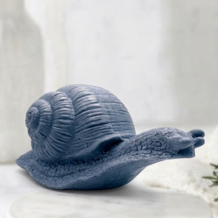 A front view of a highly detailed handmade vegan soap in a slate-grey tone, sculpted in the shape of a realistic snail with a prominent spiralled shell and a textured, leathery body, displayed on a light wooden surface.