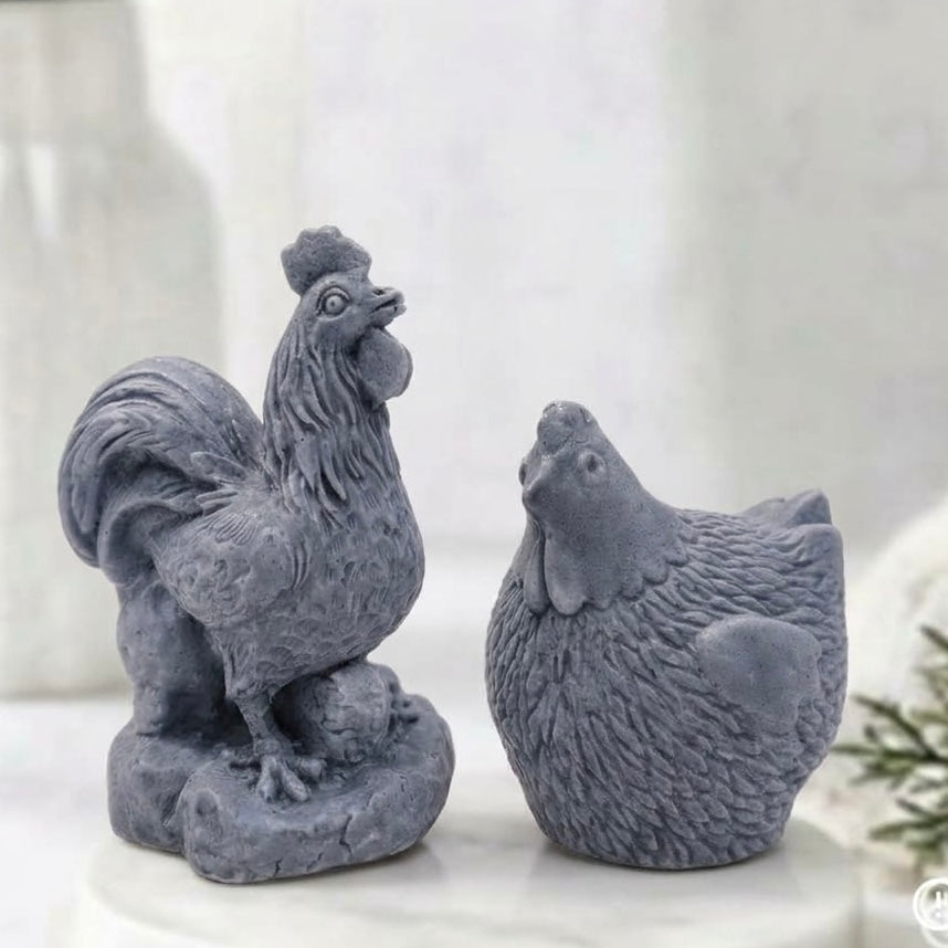 Side profile of two elegantly detailed, charcoal grey, vegan soap bars - one cockerel and one hen - resting on a white marble coaster with a bokeh background of a window and blurry greenery.