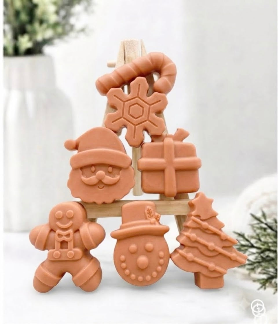 A festive collection of warm terracotta-toned mini Christmas soaps, shaped as Santa, a snowman, a Christmas tree, a gingerbread man, a present, a snowflake, and a candy cane, arranged on a mini wooden easel.