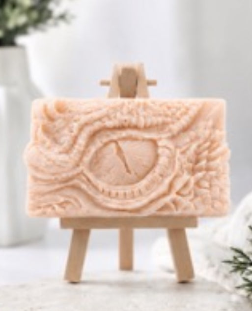 A rectangular pink French clay vegan soap bar with an embossed eye design, displayed on a small wooden easel against a soft, bright, blurred background.