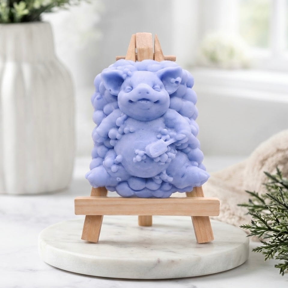 A product shot of a pig-shaped soap figurine with a bubble-cluster base, rendered in a soft blue colour, positioned full-frontal on a miniature wooden easel.
