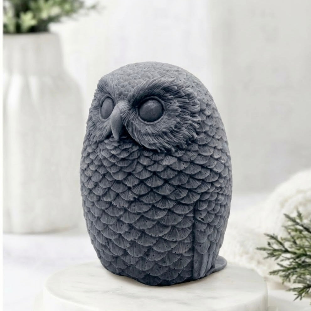 A detailed charcoal grey handmade vegan soap in the shape of an owl with intricate feather textures, displayed on a light wooden table with white flowers in the background.