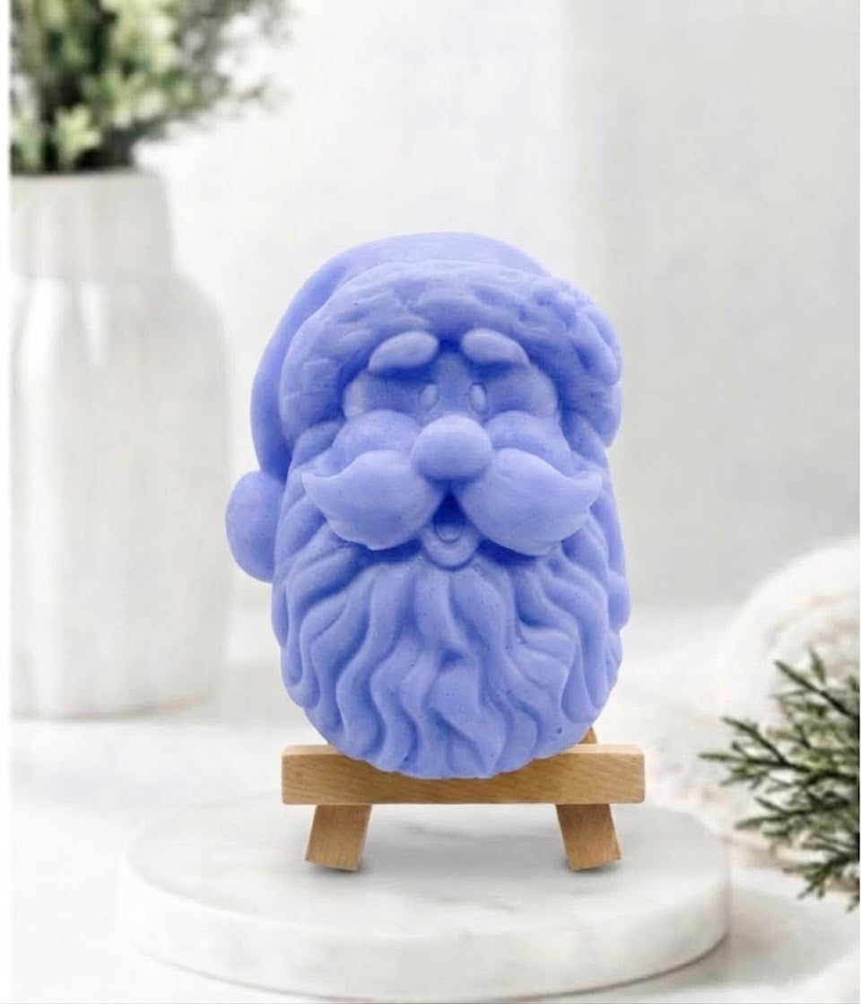 A light blue Santa Claus head soap with a detailed beard and mustache, displayed on a small wooden stand on a white marble surface with a blurred white background.