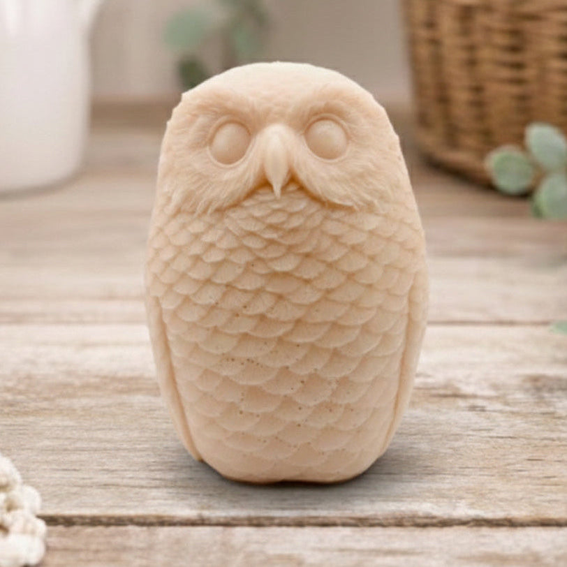 A detailed handmade vegan soap in the shape of an owl with intricate feather textures, displayed on a light wooden table with white flowers in the background.