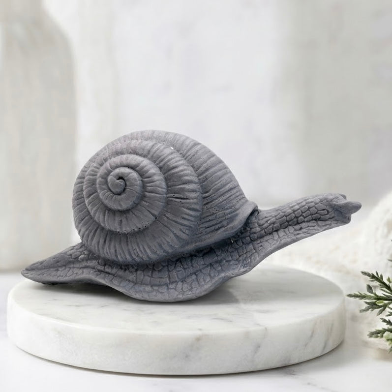 A highly detailed handmade vegan soap in a slate-grey tone, sculpted in the shape of a realistic snail with a prominent spiralled shell and a textured, leathery body, displayed on a light wooden surface.