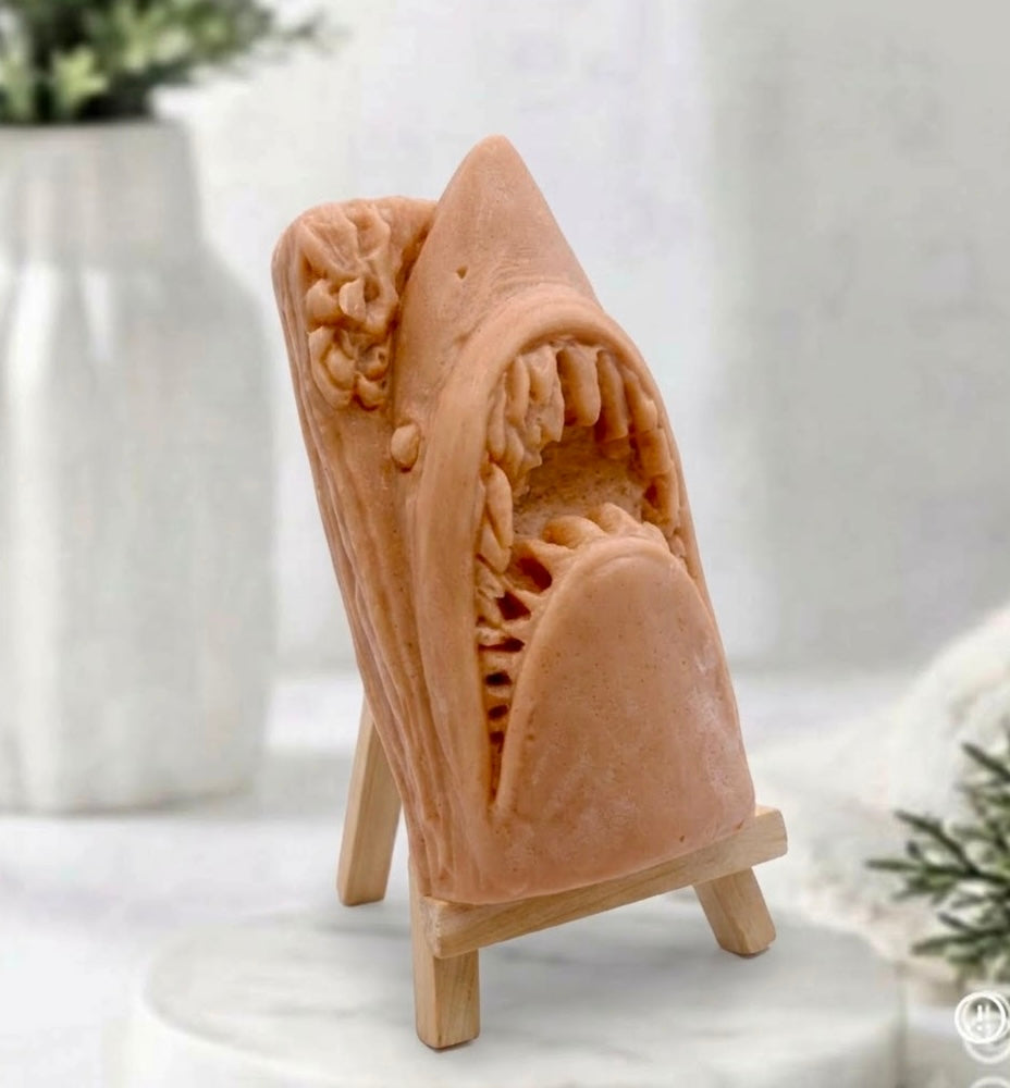 A side view of a handmade shark soap displayed on a wooden easel against a neutral background featuring a white vase and greenery.