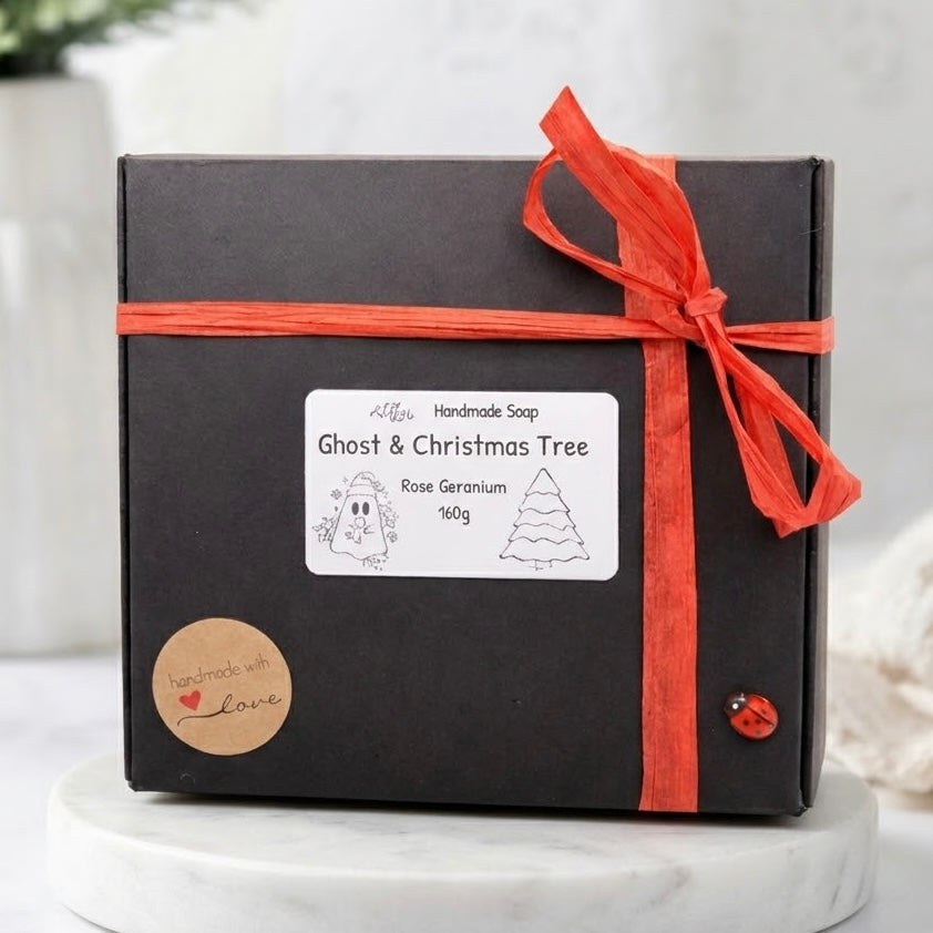 Black gift box packaging for a handmade rose geranium soap set tied with a festive red ribbon and decorated with a small ladybug accent.