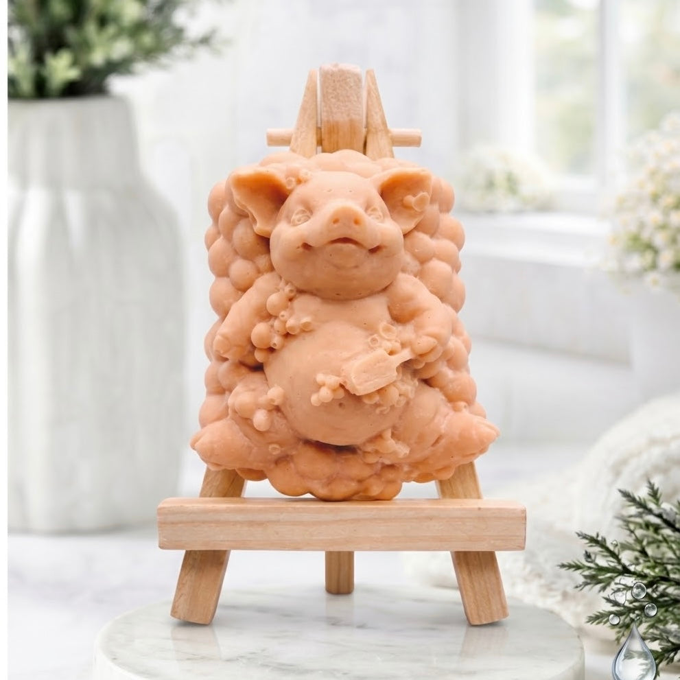 A product shot of a pig-shaped soap figurine with a bubble-cluster base, made in a terracotta colour, positioned full-frontal on a miniature wooden easel.