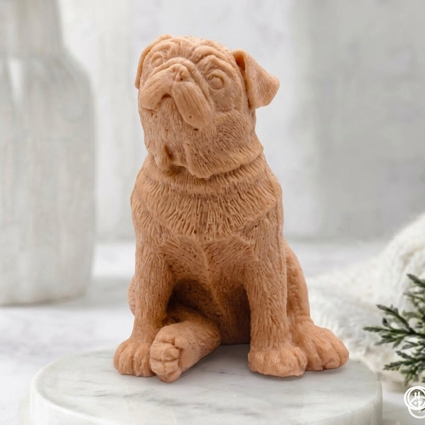 A full-body view from the front of a sitting, sculpted terracotta artisanal pug dog soap. The soap, detailed with fur texture and facial features, rests on a round marble pedestal in a calm, light-filled spa bathroom setting.