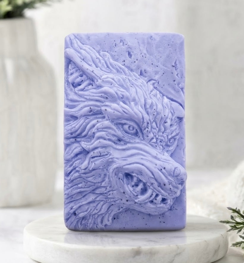 A indigo blue rectangular vegan soap bar featuring a raised relief carving of a wolf's head, displayed on a round white marble pedestal with a blurred white ceramic vase in the background.