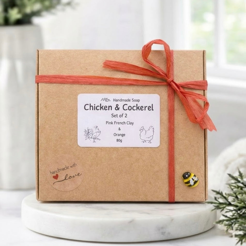 A frontal shot of a simple kraft paper box containing the 'Chicken & Cockerel' vegan soap gift set. The box is tied with a red raffia ribbon and decorated with a small bee charm. A 'handmade with love' sticker and a descriptive label detailing the set are also visible.