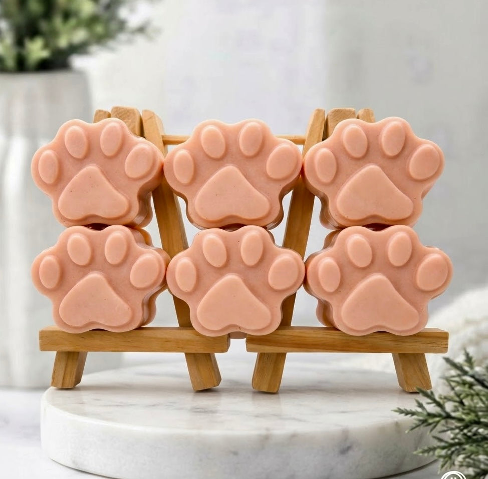 Six terracotta coloured paw print-shaped soaps on a small wooden easel, placed on a marble surface. The background includes a vase of greenery and a white towel.