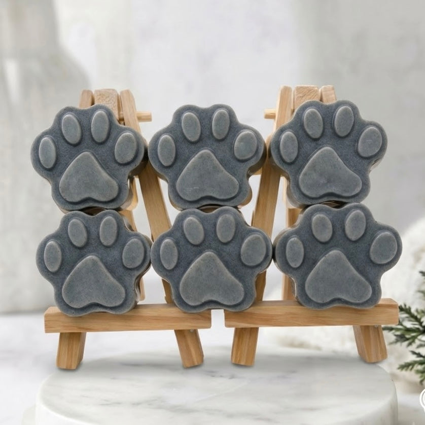 Six grey paw print-shaped soaps arranged in three stacks of two, resting on a small wooden easel on a marble surface, with a vase of eucalyptus in the background.