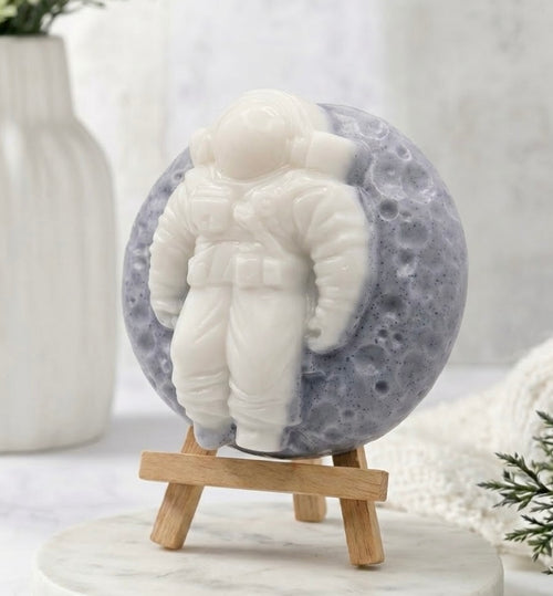 A detailed three-quarter view of the astronaut on the moon soap bar, showing the texture of the speckled blue moon base and the detailed relief of the white astronaut. The soap is displayed at an angle on the mini wooden easel.