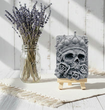 Load image into Gallery viewer, A handmade vegan soap featuring a highly detailed 3D relief of a skull wearing a jewelled crown and surrounded by roses, displayed on a wooden easel against a whimsical background.
