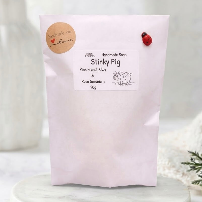 A product shot of a clean, white paper stand-up pouch packaging for soap. The pouch features a round brown paper label at the top with 'handmade with love' in cursive text. The central label identifies it as 'Handmade Soap' and 'Stinky Pig', listing ingredients 'Pink French Clay & Rose Geranium' and weight '90g', alongside a line illustration of a pig