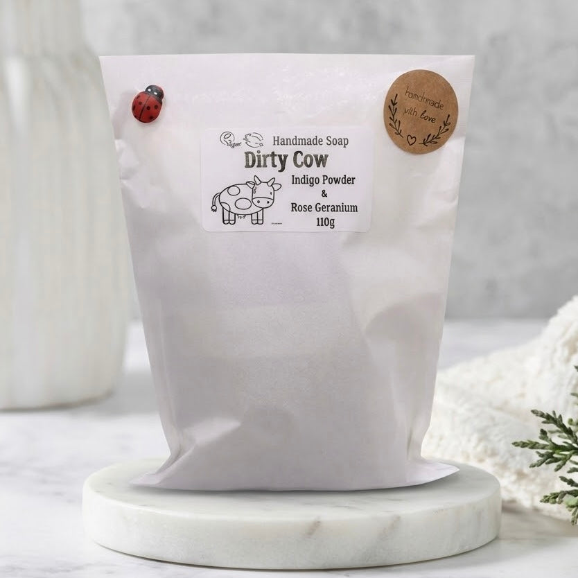Handmade vegan soap packaging for the 'Dirty Cow' bar soap with Indigo Powder and Rose Geranium, featuring a spotted cow illustration and a ladybug detail on the pouch.